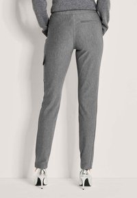 Achteraanzicht van een persoon die een nauwsluitende grijze broek met zijzakken en zilveren hoge hakken draagt, staande tegen een effen muur.