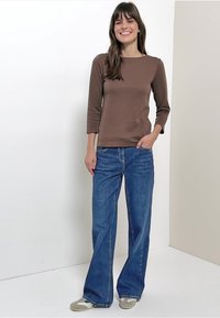 Brązowy top z długim rękawem i okrągłym dekoltem, zestawiony z niebieskimi dzwonowatymi dżinsami. Miękka tkanina i luźny krój. Stylizację dopełniają jasne sportowe buty.
