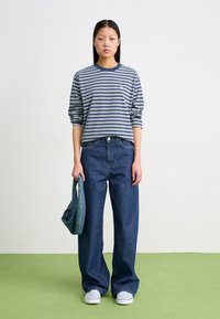 Camisa de algodão de manga longa, às riscas azul-marinho e brancas, combinada com uns jeans de ganga escuros e soltos, e ténis brancos. A segurar uma pequena bolsa de ganga.