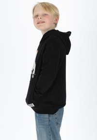 Black hoodie with front graphic design, kangaroo pocket, ribbed cuffs, and hem. Worn over light blue jeans, model posed sideways.