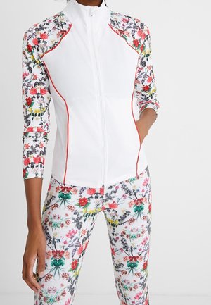 Biała kurtka na zamek z rękawami i panelami bocznymi w kwiatowy wzór, noszona z dopasowanymi legginsami w ten sam kwiatowy wzór, zawierającym czerwone, zielone, żółte i czarne kwiaty.