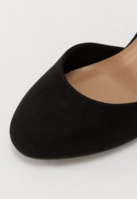 Svart mocka högklackad sko med rund tå och djupt inskuren fram. Slen textur med en beige innersula synlig inom öppningen.