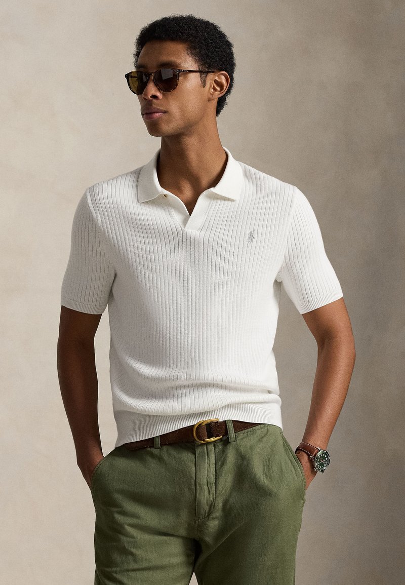 White ribbed short-sleeve polo shirt with a collar, worn with olive green trousers, featuring a brown belt and a silver watch.