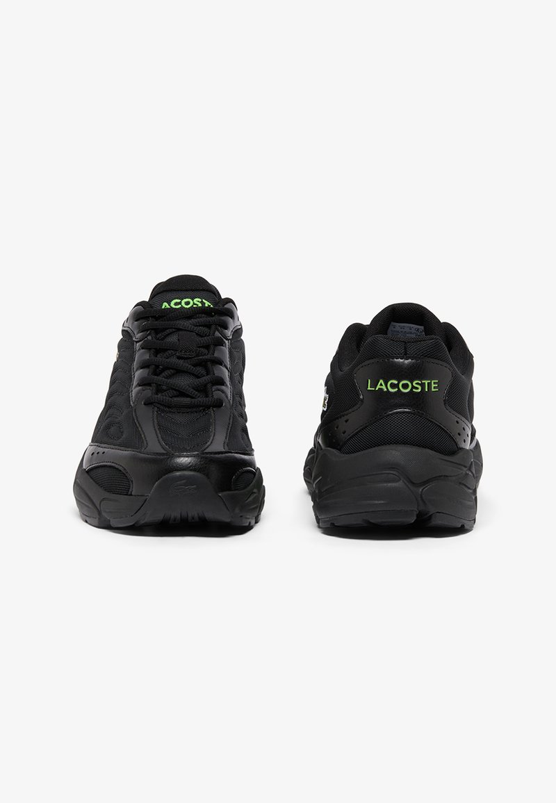 Paire de chaussures de sport noires montrant la vue avant de la chaussure gauche et la vue arrière de la chaussure droite avec le logo "LACOSTE" vert sur la languette et le talon.