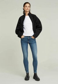 Μαύρη μπόμπερ τζάκετ, λευκό T-shirt, μπλε skinny τζιν και μαύρες μποτάκια. Λείο υλικό, εφαρμοστή γραμμή και χαλαρά μανίκια στο τζάκετ.