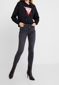 Sudadera corta negra con logotipo rojo y blanco, combinada con jeans ajustados gris oscuro y botas negras de tobillo con punta afilada. Texturas suaves en todo el conjunto.