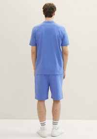 Světle modrá bavlněná polo tričko a odpovídající šortky, s volným střihem, krátkými rukávy a bočními rozparky na lemu. Viditelné bílé tenisky a ponožky.