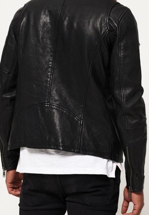 Person iført en sort læderjakke med lynlåse på ærmerne over en hvid skjorte og sorte bukser, set bagfra.