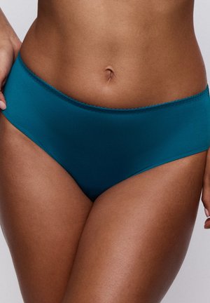 Vrouw draagt turquoise naadloos ondergoed, waarbij de taille en bovenbenen zichtbaar zijn tegen een effen grijze achtergrond.