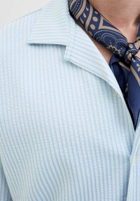 Lichtblauw geklede shirt met verticale witte strepen, met een getextureerde stof en knoopsluiting. Een marineblauwe gedetailleerde sjaal is om de hals geknoopt.