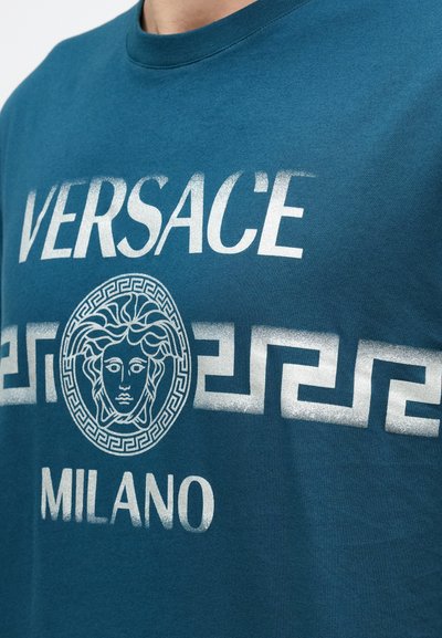 Tirkīzzils kokvilnas T-krekls ar baltu grafiku, uz kuras redzams uzraksts "VERSACE" un "MILANO", kā arī apaļa Medūzas emblēma, ko ieskauj grieķu atslēgas raksts.