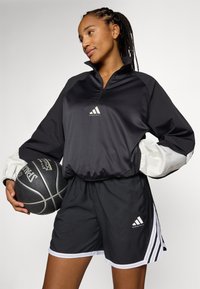Svart athletic pullover med hvite detaljer, med høy krage og logo, kombinert med svarte shorts med hvit kant og striper, og holder en basketball.