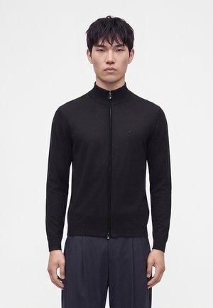 Homme aux cheveux courts et noirs portant un pull zippé noir et un pantalon sombre, debout devant un fond blanc uni.