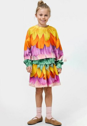 Petite fille souriante, portant une robe à motif de pétales de fleurs colorés avec des manches longues, des chaussettes roses et des chaussures plates marron.