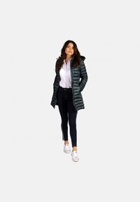 Veste matelassée vert foncé avec capuche et motif vertical de piqûres, portée sur un polo blanc et associée à un jean noir et des baskets blanches.