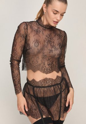 Frau trägt schwarzen transparenten Spitzen-Langarm-Crop-Top und passenden Rock mit Strapshaltern über schwarzer Unterwäsche, schaut nach unten.