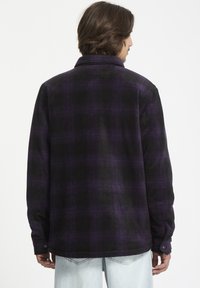 Camisa de cuadros en tonos morados y negros, con cuello abotonado, mangas largas y una textura suave, que presenta un corte recto y puños ajustables.