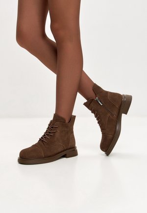 Bottines en daim marron avec lacets et fermetures éclair latérales portées sur des jambes croisées sur fond blanc.