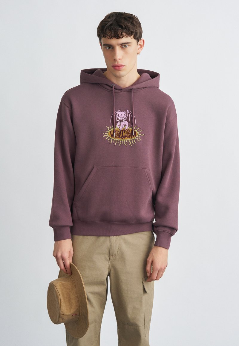 Sweat à capuche marron avec une poche avant, orné d'un motif graphique coloré. Porté avec un pantalon kaki et tenant un chapeau beige.
