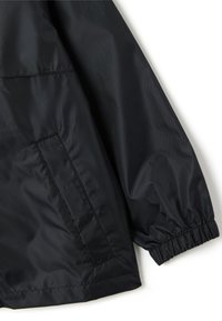 Manche de veste en nylon noir avec poignet élastique et détail de poche latérale sur un fond blanc.