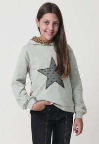 Sudadera verde claro con un diseño de estrella negra y lunares, con forro de capucha de estampado de leopardo y puños elásticos. Jeans negros con botones dorados.
