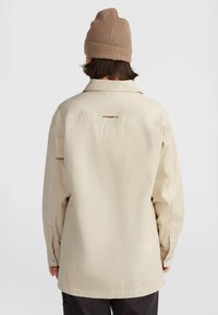 Camisa de algodón beige con mangas largas, collar y corte recto; presenta un sutil logo en la parte superior del canesú y puños abotonados.