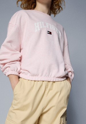 Personne portant un sweat-shirt rose clair avec le texte et le logo "HILFIGER", associé à un pantalon cargo beige, les mains dans les poches, sur fond gris.