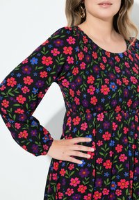 Czarna sukienka w kwiaty z intensywnymi różowymi, niebieskimi i purpurowymi kwiatami oraz zielonymi liśćmi, z marszczonym gorsetem i długimi rękawami. Gładka tekstura tkaniny.