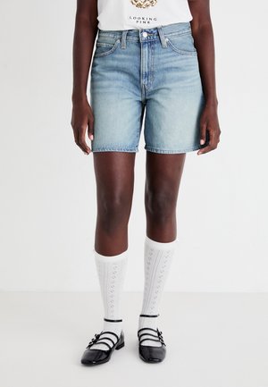 Person iført lyseblå denimshorts, hvide knæstrømper med mønster, sorte remsko og en hvid t-shirt med grafik.