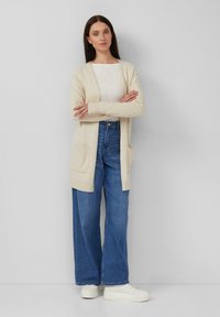 Donna in piedi con le braccia incrociate, indossa un cardigan beige, una camicia bianca, jeans larghi blu e sneakers platform bianche, di fronte a un muro grigio.