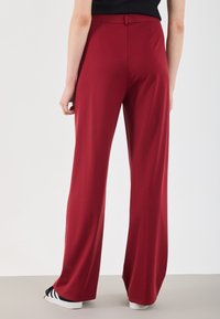 Red wide-leg trousers in a smooth, stretchy fabric with a fitted waistband, featuring a subtle sheen and clean finish at hem. White sneakers visible.