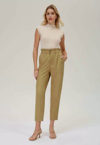 Beige ärmelloses Oberteil und hochtaillierte, schmal zulaufende olivgrüne Hose mit einer subtilen Textur. Dünne, hautfarbene Sandalen betonen den Look.