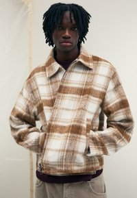 Hnedo-krémový kostkovaný kabát vyrobený z mäkkého, štruktúrovaného materiálu; má golier, zips vpredu a bočné vrecká. Nosený cez tmavý top.