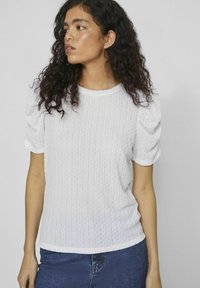 Haut blanc texturé à manches courtes avec un col rond et des manches bouffantes froncées, porté avec une jupe en denim bleue.
