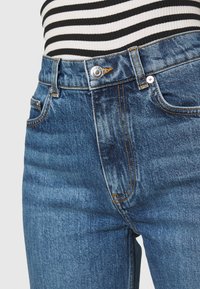 Gros plan sur un jean en denim bleu avec des coutures orange et un bouton argenté, porté avec un haut à rayures noires et blanches.
