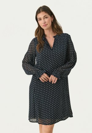 Laivastonsininen mekko, jossa on valkoisia pallokuvioita, pitkät läpinäkyvät hihat, viistetty pääntie ja rento malli, joka ulottuu polven ylle.