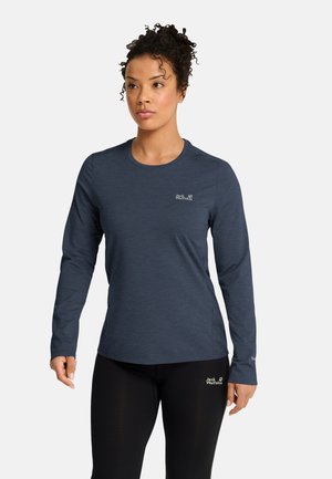Frau mit lockigem Haar, die ein dunkles Langarmshirt und schwarze Hosen mit Jack Wolfskin-Logos auf beiden Kleidungsstücken trägt.