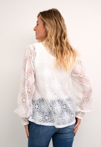 Blusa bianca in pizzo con motivi floreali, maniche lunghe a sbuffo e orlo leggermente curvo. Indossata sopra jeans di denim blu, mostrando dettagli intricati.