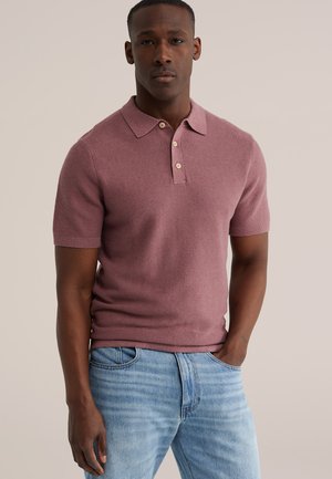 Homme portant un polo à manches courtes mauve avec des boutons et un jean bleu clair, debout avec une main dans la poche devant un fond uni.
