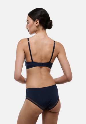 Femme aux cheveux foncés attachés en chignon, portant un soutien-gorge bleu marine et une culotte assortie, vue de dos sur un fond clair uni.