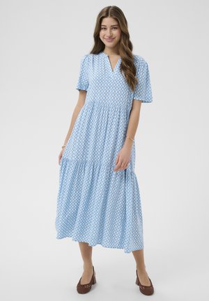 Jeune femme debout, souriante, portant une robe midi bleu clair à motif de cercles blancs et des chaussures à talons carrés en tissage marron.