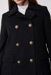 Manteau noir croisé en laine, avec des boutons en relief doré représentant un lion, de larges rabats et des poches latérales.