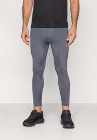 Vyras dėvi aptemptas pilkas sportines kelnes ir juodus bėgimo batelius, stovi priešais šviesią vientisą foną.