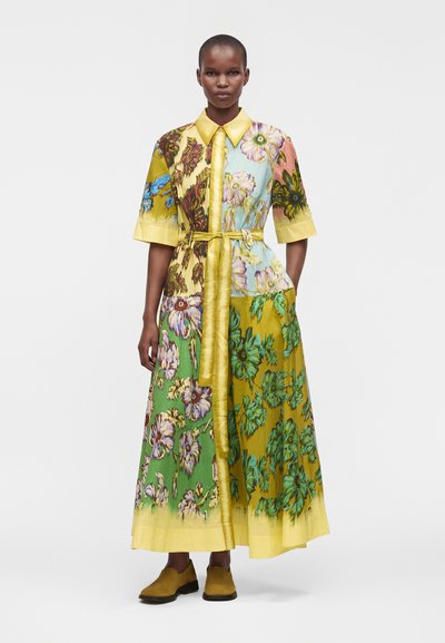 Femme portant une longue robe patchwork florale multicolore avec un col et une ceinture jaunes, debout les mains dans les poches et des chaussures marron.