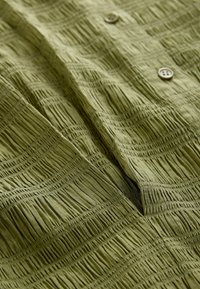 Rakennepintainen vihreä kangas, jossa on ruudukkomalli ja kerättyjä yksityiskohtia. Kaksi ruskeaa nappia näkyvissä, muodostaen kerroksellisen ilmeen.