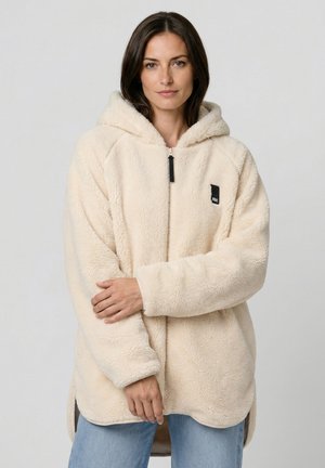 Frau trägt einen cremefarbenen Fleece-Hoodie mit Reißverschluss und schwarzem Logo-Patch, kombiniert mit hellblauen Jeans, und steht vor einem einfarbigen Hintergrund.