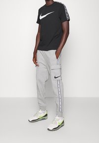 Svart Nike T-shirt med vit logotyp, grå cargobyxor med sidfickor och vita sidosträck, samt vita sneakers med gröna accenter.