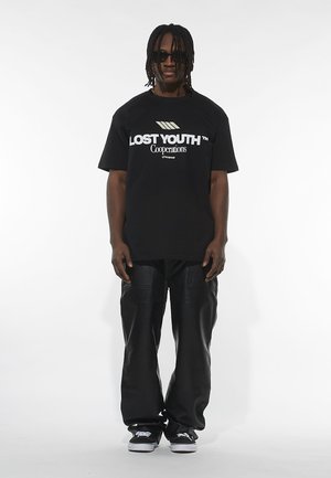 Svart bomull t-skjorte med hvit tekst "LOST YOUTH™ Cooperations," kombinert med skinnbukser og svarte sneakers. Modellen har på seg solbriller.