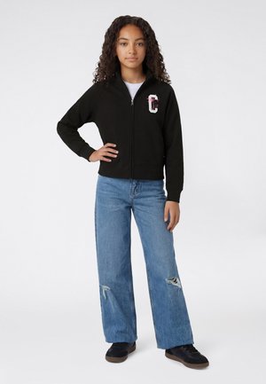 Mädchen mit lockigem Haar, das eine schwarze Jacke mit Reißverschluss und einem pinkfarbenen Buchstaben "C" als Aufdruck, zerrissene Jeans mit weitem Bein in Blau und schwarze Sneaker trägt und steht.