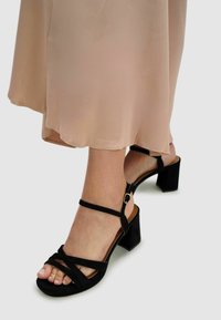 Black suede heeled sandals featuring a multi-strap design, ankle straps with a gold buckle, and a textured block heel.
